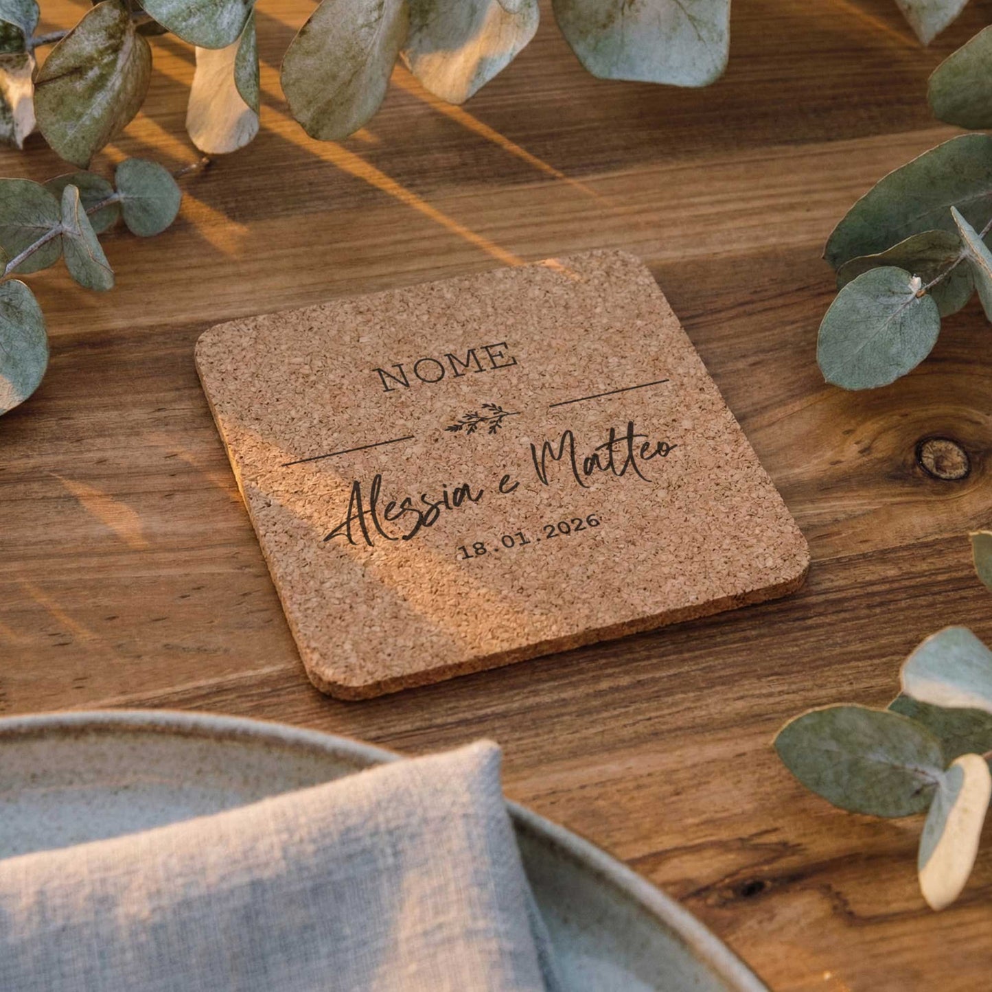 Segnaposto matrimonio personalizzato in sughero quadrato inciso, appoggiato su tavolo in legno con decorazioni botaniche ed eucalipto.