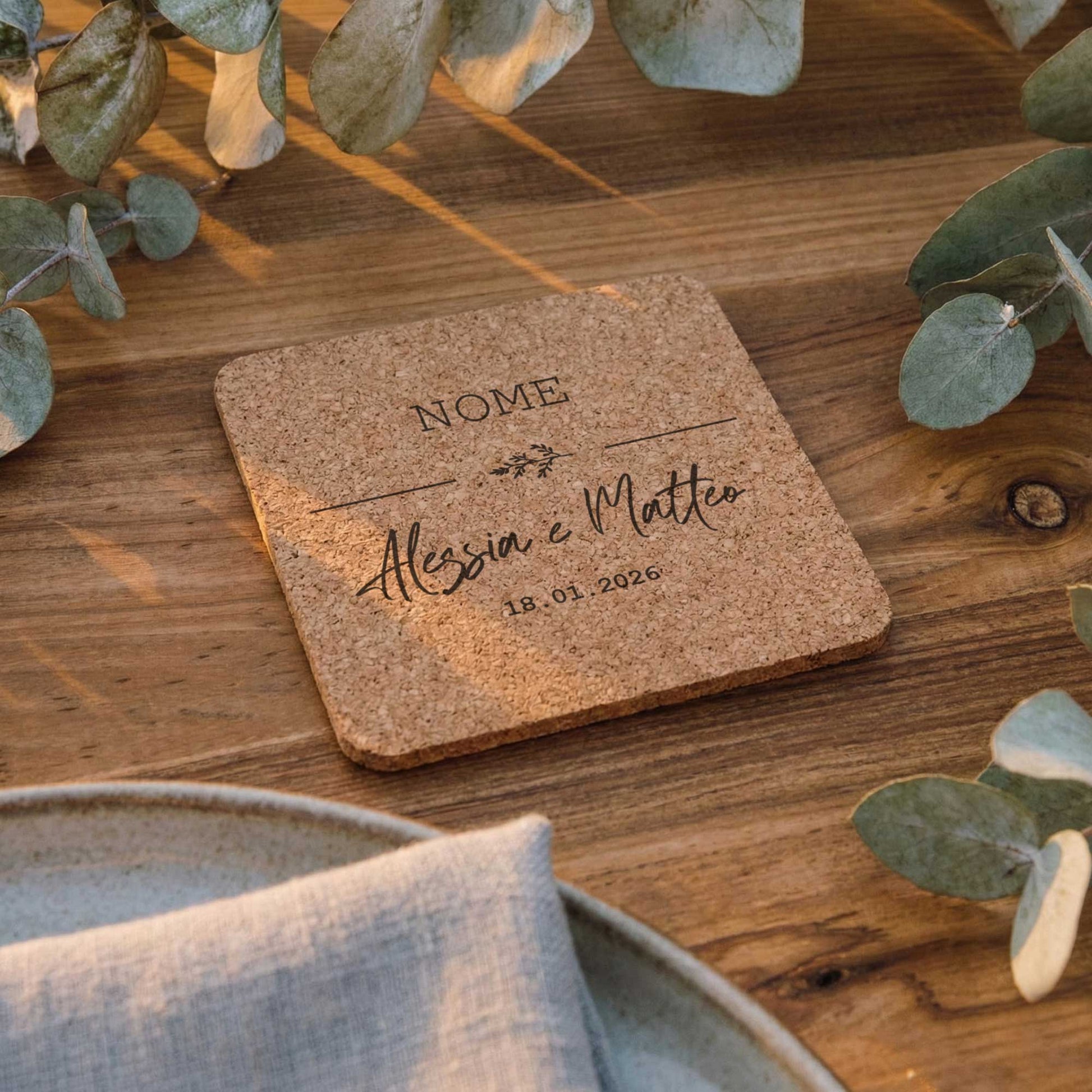 Segnaposto matrimonio personalizzato in sughero quadrato inciso, appoggiato su tavolo in legno con decorazioni botaniche ed eucalipto.